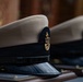 The image shows two military or naval dress uniform caps placed on a wooden surface. The caps feature a white/khaki crown, black band, black patent leather visor, and gold anchor insignia on the front. The composition is a close-up shot with shallow depth of field, keeping the foreground cap in sharp focus.