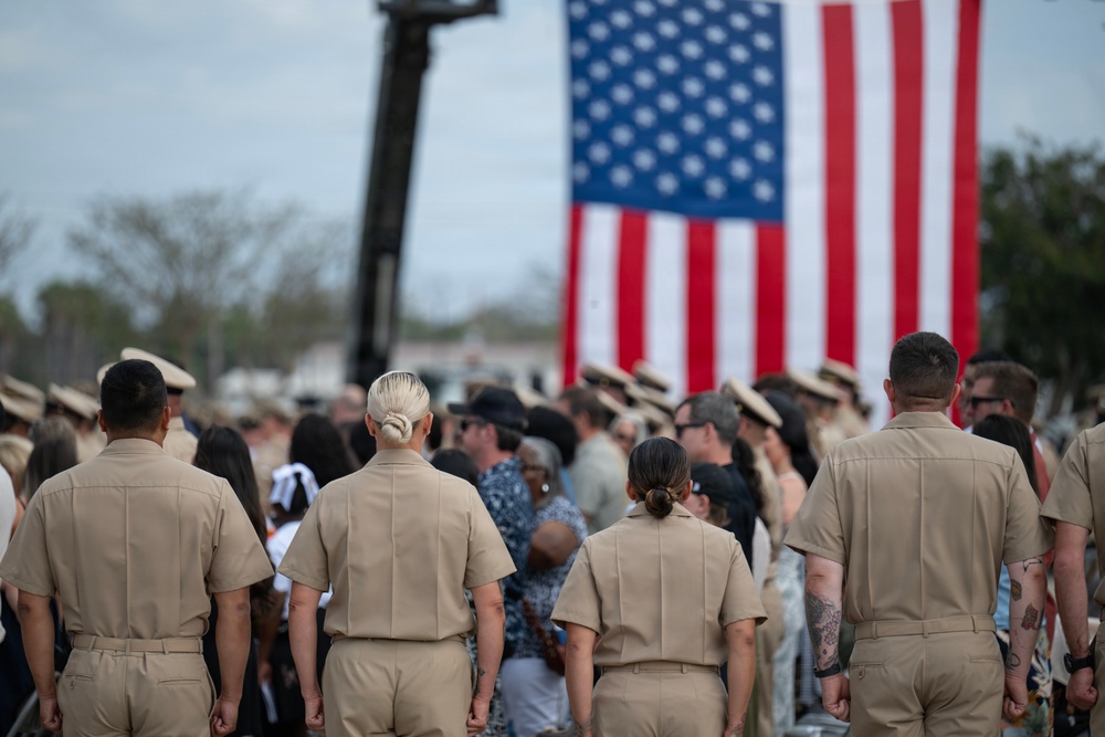 A group of U.S. Navy personnel in tan/khaki service uniforms are gathered at what appears to be a military ceremony or formation, facing toward a large American flag displayed in the background. The scene includes both uniformed military members and civilians, suggesting a public or commemorative event. The large flag is prominently displayed, likely suspended from a crane or aerial lift vehicle visible in the background.