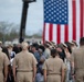 A group of U.S. Navy personnel in tan/khaki service uniforms are gathered at what appears to be a military ceremony or formation, facing toward a large American flag displayed in the background. The scene includes both uniformed military members and civilians, suggesting a public or commemorative event. The large flag is prominently displayed, likely suspended from a crane or aerial lift vehicle visible in the background.