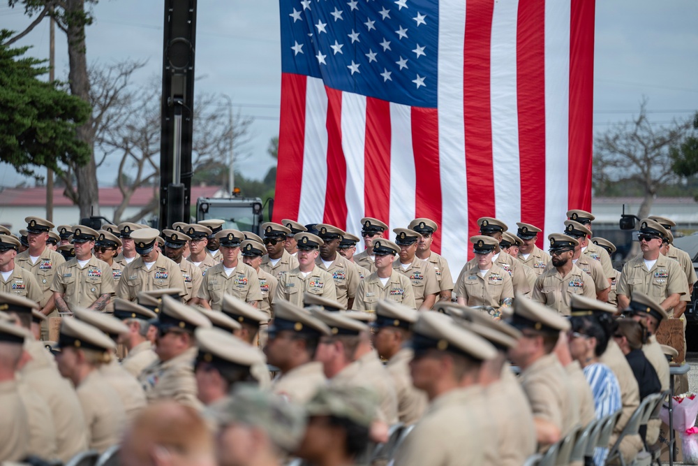 A large formation of U.S. Navy personnel in khaki service uniforms are assembled at an outdoor military ceremony. A large American flag is prominently displayed in the background, suggesting a formal event such as a change of command, promotion ceremony, or commissioning. Personnel are arranged in rows, with a standing rank visible behind seated attendees in the foreground.