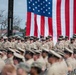 A large formation of U.S. Navy personnel in khaki service uniforms are assembled at an outdoor military ceremony. A large American flag is prominently displayed in the background, suggesting a formal event such as a change of command, promotion ceremony, or commissioning. Personnel are arranged in rows, with a standing rank visible behind seated attendees in the foreground.