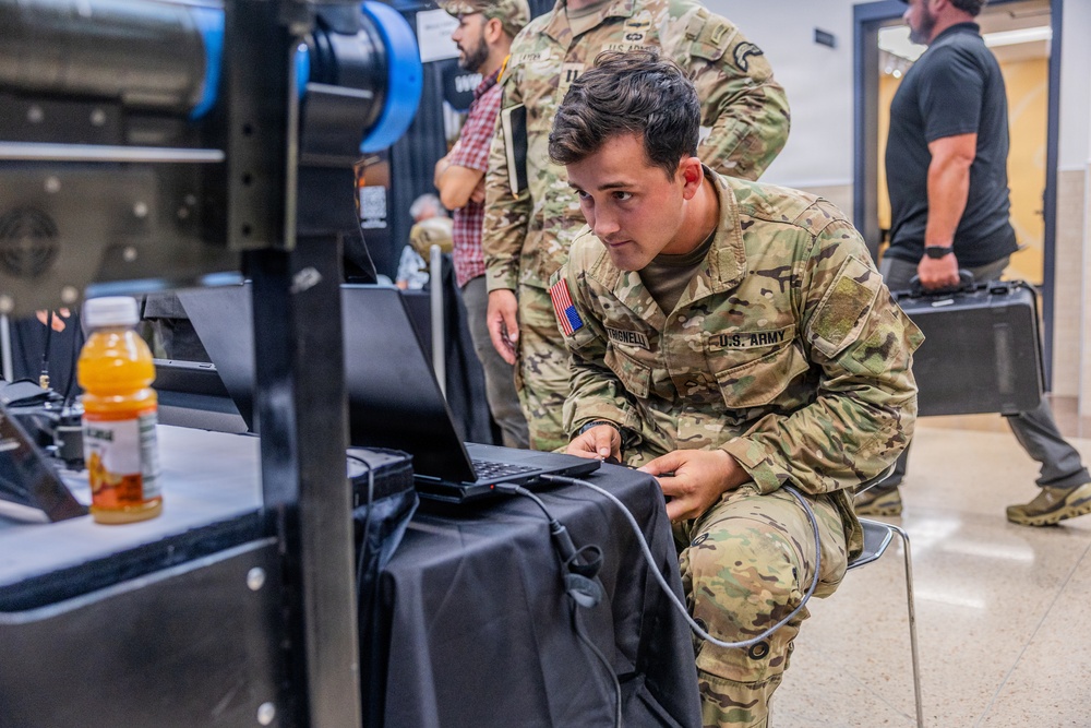 A U.S. Army soldier in MultiCam/OCP camouflage uniform is crouched down working on a laptop computer at what appears to be a military technology demonstration or expo event. The soldier's name tape reads what appears to be 'TRIGNELLI' and the branch tape reads 'U.S. ARMY'. Several other military personnel and civilians are visible in the background of the indoor venue.