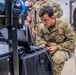 A U.S. Army soldier in MultiCam/OCP camouflage uniform is crouched down working on a laptop computer at what appears to be a military technology demonstration or expo event. The soldier's name tape reads what appears to be 'TRIGNELLI' and the branch tape reads 'U.S. ARMY'. Several other military personnel and civilians are visible in the background of the indoor venue.