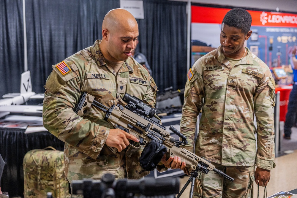 Two U.S. Army soldiers are examining a rifle at what appears to be a military equipment exhibition or trade show. The soldier in the foreground, identified by name tape as 'PADILLA,' is handling and inspecting the weapon while soldier 'SWAINSON' observes. The background shows vendor booths, including one for 'Leonardo,' suggesting a defense industry expo environment.