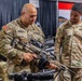 Two U.S. Army soldiers are examining a rifle at what appears to be a military equipment exhibition or trade show. The soldier in the foreground, identified by name tape as 'PADILLA,' is handling and inspecting the weapon while soldier 'SWAINSON' observes. The background shows vendor booths, including one for 'Leonardo,' suggesting a defense industry expo environment.