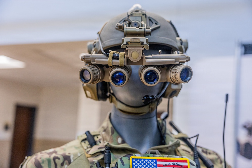 A military mannequin is displayed wearing advanced tactical gear, including a multi-lens night vision goggle system mounted on a combat helmet. The mannequin is dressed in MultiCam/OCP camouflage uniform with visible patches including a US flag and a patch reading 'nous défions'. The display appears to be in an indoor exhibition or briefing room setting.