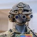 A military mannequin is displayed wearing advanced tactical gear, including a multi-lens night vision goggle system mounted on a combat helmet. The mannequin is dressed in MultiCam/OCP camouflage uniform with visible patches including a US flag and a patch reading 'nous défions'. The display appears to be in an indoor exhibition or briefing room setting.