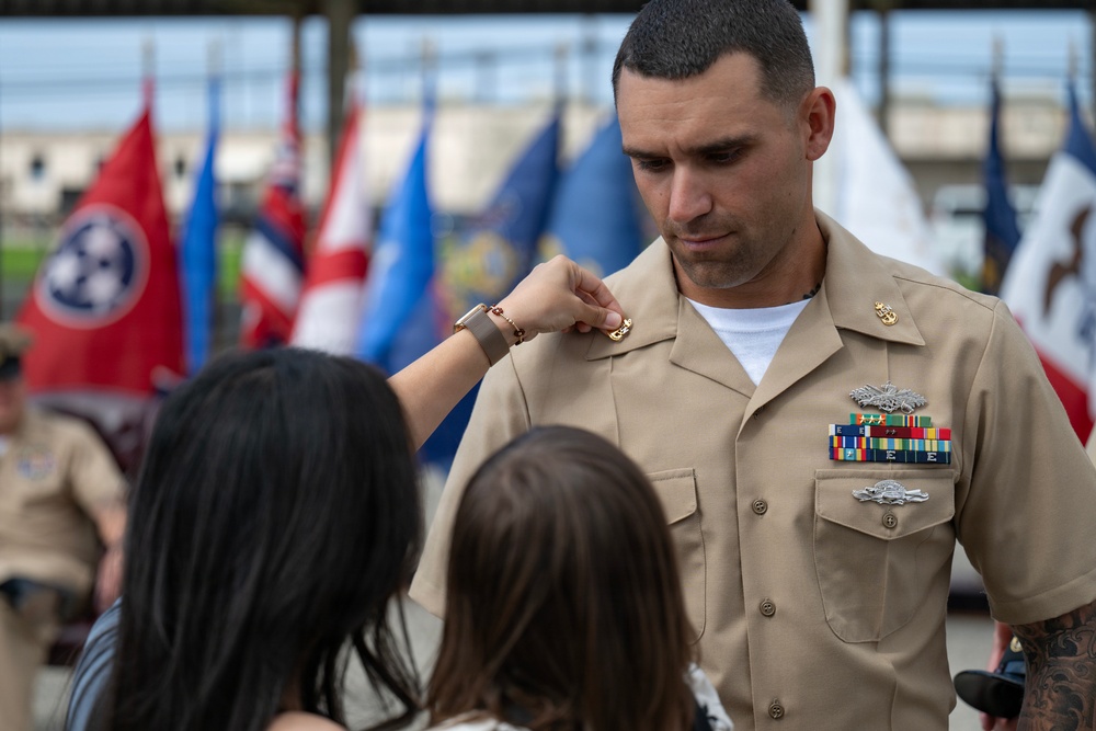 A U.S. Navy sailor in khaki service uniform is having a rank insignia or device pinned onto his collar by what appears to be a family member during a promotion or advancement ceremony. Multiple national and state flags are visible in the background, indicating a formal military ceremony setting. The sailor displays several service ribbons, qualification badges, and collar devices on his uniform.