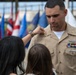 A U.S. Navy sailor in khaki service uniform is having a rank insignia or device pinned onto his collar by what appears to be a family member during a promotion or advancement ceremony. Multiple national and state flags are visible in the background, indicating a formal military ceremony setting. The sailor displays several service ribbons, qualification badges, and collar devices on his uniform.