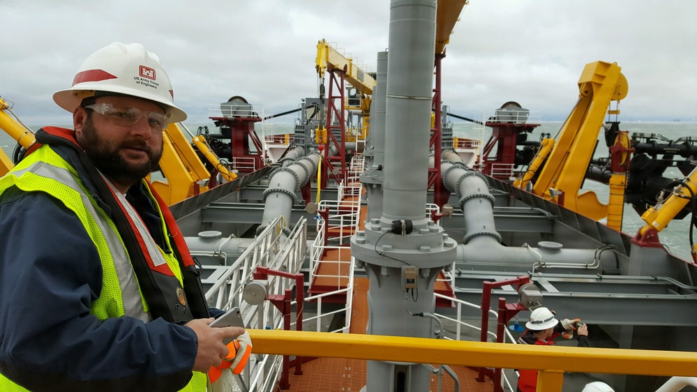 A man wearing a US Army Corps of Engineers hard hat, safety glasses, high-visibility vest, and personal flotation device stands on the deck of a large dredging vessel. The ship features extensive industrial piping, yellow cranes, and red structural supports, with open water visible in the background. At least two other workers in safety gear are visible in the background on the lower deck.
