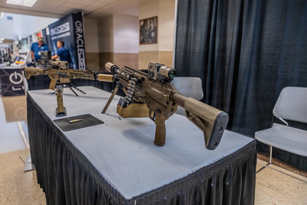 The image shows a firearms display at what appears to be a trade show or military/law enforcement exhibition. Multiple rifles in flat dark earth (FDE/tan) finish are displayed on a skirted table, with at least one featuring a bipod, optics, and stacked magazines. A banner reading 'ORACLE' is visible in the background along with booth personnel.