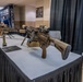 The image shows a firearms display at what appears to be a trade show or military/law enforcement exhibition. Multiple rifles in flat dark earth (FDE/tan) finish are displayed on a skirted table, with at least one featuring a bipod, optics, and stacked magazines. A banner reading 'ORACLE' is visible in the background along with booth personnel.