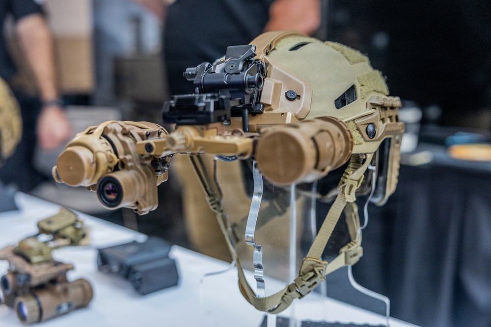 A tan/coyote-colored tactical military helmet is displayed on a clear acrylic stand on a white table, likely at a defense or military equipment exhibition. The helmet is equipped with a multi-tube night vision goggle (NVG) system mounted on a front bracket arm. Additional military optical equipment is visible in the background on the same display table.