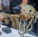 A tan/coyote-colored tactical military helmet is displayed on a clear acrylic stand on a white table, likely at a defense or military equipment exhibition. The helmet is equipped with a multi-tube night vision goggle (NVG) system mounted on a front bracket arm. Additional military optical equipment is visible in the background on the same display table.