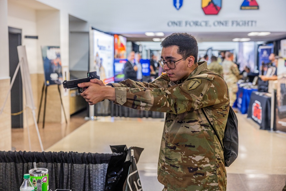 A soldier in US Army OCP (Operational Camouflage Pattern) uniform is handling and aiming a pistol at what appears to be a military expo or trade show. The event features multiple vendor booths and displays in the background, with other uniformed personnel visible. A banner reading 'ONE FORCE, ONE FIGHT!' is visible overhead, along with military unit insignia patches.