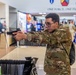 A soldier in US Army OCP (Operational Camouflage Pattern) uniform is handling and aiming a pistol at what appears to be a military expo or trade show. The event features multiple vendor booths and displays in the background, with other uniformed personnel visible. A banner reading 'ONE FORCE, ONE FIGHT!' is visible overhead, along with military unit insignia patches.