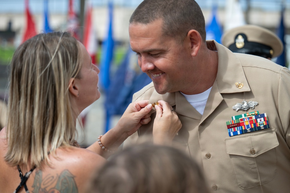 A woman is pinning an insignia or device onto the collar of a smiling U.S. Navy enlisted sailor in khaki service uniform during what appears to be a promotion or advancement ceremony. The sailor displays multiple service ribbons, a warfare device, and collar insignia on his uniform. The outdoor ceremony setting includes military flags visible in the blurred background.