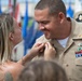 A woman is pinning an insignia or device onto the collar of a smiling U.S. Navy enlisted sailor in khaki service uniform during what appears to be a promotion or advancement ceremony. The sailor displays multiple service ribbons, a warfare device, and collar insignia on his uniform. The outdoor ceremony setting includes military flags visible in the blurred background.