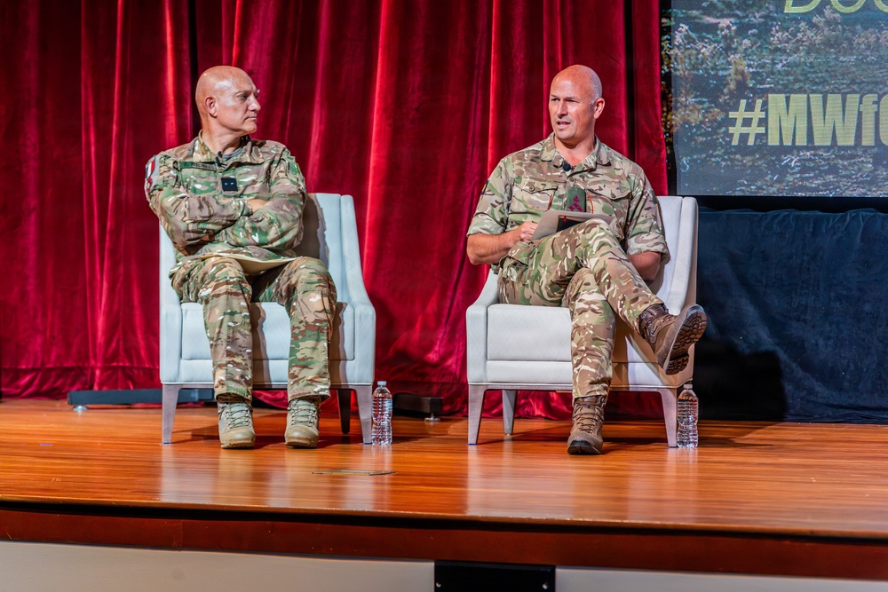 Two military personnel in camouflage uniforms are seated on white chairs on a wooden stage during what appears to be a panel discussion or speaking event. The person on the right is actively speaking and holding papers, while the person on the left listens with arms crossed. A red curtain backdrop and a screen displaying '#MWf...' are visible behind them.