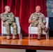 Two military personnel in camouflage uniforms are seated on white chairs on a wooden stage during what appears to be a panel discussion or speaking event. The person on the right is actively speaking and holding papers, while the person on the left listens with arms crossed. A red curtain backdrop and a screen displaying '#MWf...' are visible behind them.