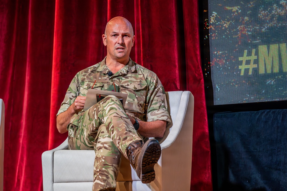 A bald male individual wearing British MTP (Multi-Terrain Pattern) camouflage military uniform is seated in a white modern chair on what appears to be a stage or panel discussion setting. He is holding a tablet or document and appears to be speaking or presenting. The backdrop includes red velvet curtains and a digital display screen showing a hashtag partially visible as '#MY...'.