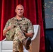 A bald male individual wearing British MTP (Multi-Terrain Pattern) camouflage military uniform is seated in a white modern chair on what appears to be a stage or panel discussion setting. He is holding a tablet or document and appears to be speaking or presenting. The backdrop includes red velvet curtains and a digital display screen showing a hashtag partially visible as '#MY...'.