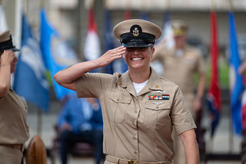 A female U.S. Navy Chief Petty Officer in Service Khaki uniform is rendering a salute with a broad smile during what appears to be a formal military ceremony. She is wearing a combination cover (cap) with a gold anchor device, service ribbons, aviation warfare insignia, and collar devices. The background shows multiple military flags and other uniformed personnel in a blurred outdoor setting.