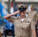 A female U.S. Navy Chief Petty Officer in Service Khaki uniform is rendering a salute with a broad smile during what appears to be a formal military ceremony. She is wearing a combination cover (cap) with a gold anchor device, service ribbons, aviation warfare insignia, and collar devices. The background shows multiple military flags and other uniformed personnel in a blurred outdoor setting.
