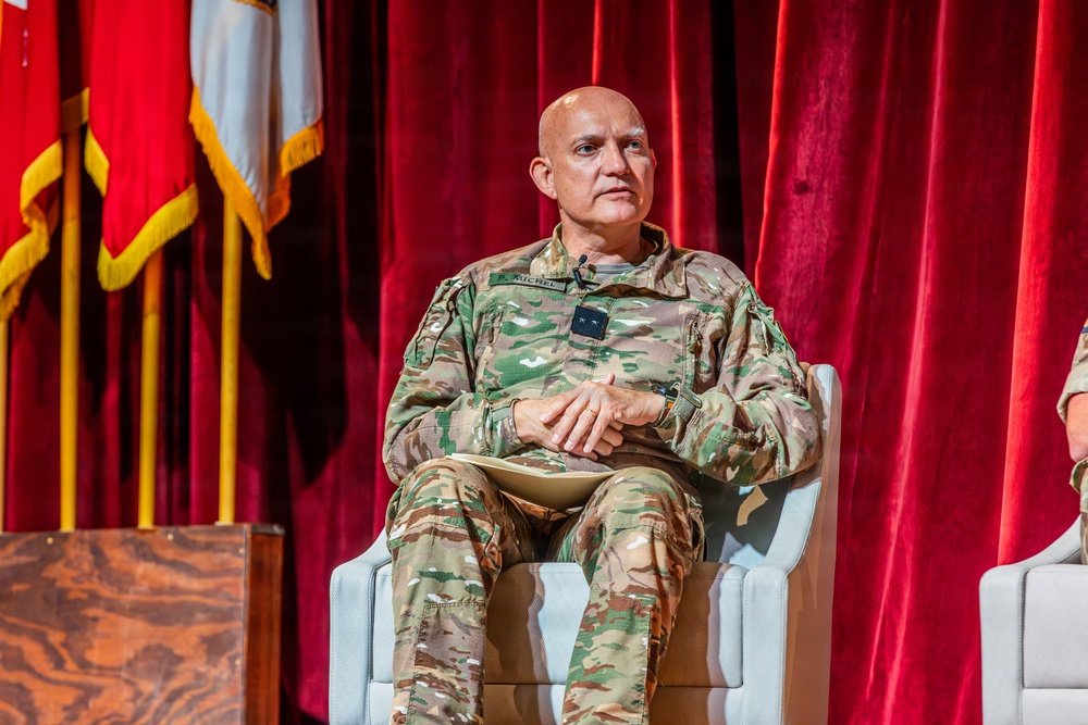 A military officer in U.S. Army OCP (Operational Camouflage Pattern) uniform is seated in a white chair on what appears to be a formal stage or auditorium setting. The officer has a name tape reading 'MICHEL' and rank insignia visible on the chest. The setting suggests a formal military briefing, panel discussion, or ceremony.