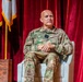 A military officer in U.S. Army OCP (Operational Camouflage Pattern) uniform is seated in a white chair on what appears to be a formal stage or auditorium setting. The officer has a name tape reading 'MICHEL' and rank insignia visible on the chest. The setting suggests a formal military briefing, panel discussion, or ceremony.