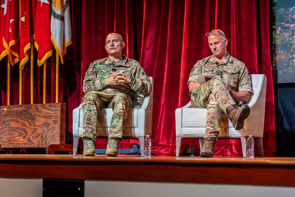 Two military personnel in camouflage uniforms are seated in white armchairs on a stage during what appears to be a formal panel discussion or briefing. The setting includes red curtains, military flags with gold fringe in the background, and a wooden podium to the left. One individual appears to be speaking while the other reviews notes on a tablet or document.