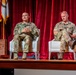 Two military personnel in camouflage uniforms are seated in white armchairs on a stage during what appears to be a formal panel discussion or briefing. The setting includes red curtains, military flags with gold fringe in the background, and a wooden podium to the left. One individual appears to be speaking while the other reviews notes on a tablet or document.