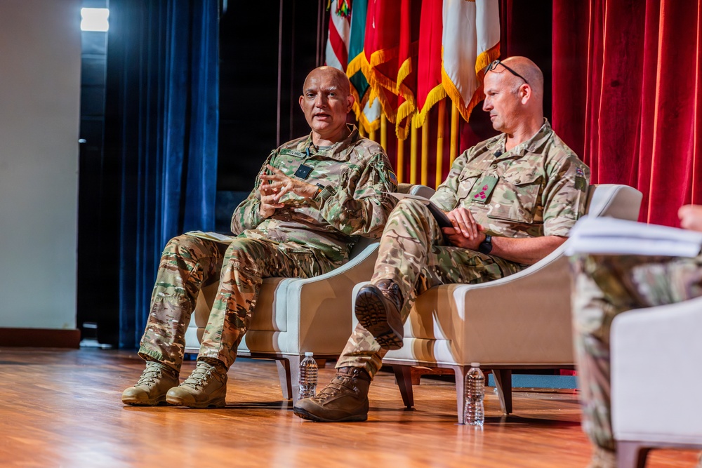 Two military personnel in camouflage uniforms (OCP/MultiCam pattern) are seated in white chairs on a stage during what appears to be a formal panel discussion or briefing. The individual on the left is actively speaking and gesturing, while the one on the right listens and holds a tablet or folder. The setting includes military flags, red and blue curtains, and a wooden stage floor.