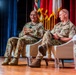 Two military personnel in camouflage uniforms (OCP/MultiCam pattern) are seated in white chairs on a stage during what appears to be a formal panel discussion or briefing. The individual on the left is actively speaking and gesturing, while the one on the right listens and holds a tablet or folder. The setting includes military flags, red and blue curtains, and a wooden stage floor.
