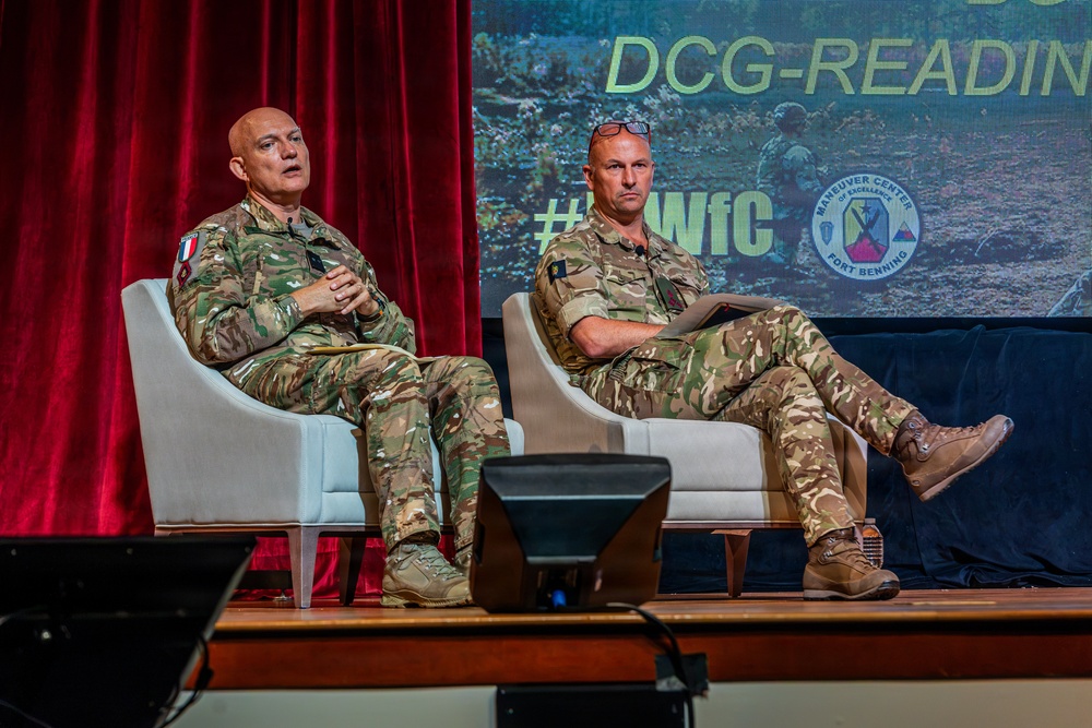 Two military personnel in MultiCam/MTP camouflage uniforms are seated in white chairs on a stage, appearing to participate in a panel discussion or briefing. The backdrop displays 'DCG-READING' text and the Maneuver Center of Excellence, Fort Benning logo, along with a hashtag '#WfC'. The setting appears to be a formal military conference or symposium.