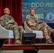 Two military personnel in MultiCam/MTP camouflage uniforms are seated in white chairs on a stage, appearing to participate in a panel discussion or briefing. The backdrop displays 'DCG-READING' text and the Maneuver Center of Excellence, Fort Benning logo, along with a hashtag '#WfC'. The setting appears to be a formal military conference or symposium.