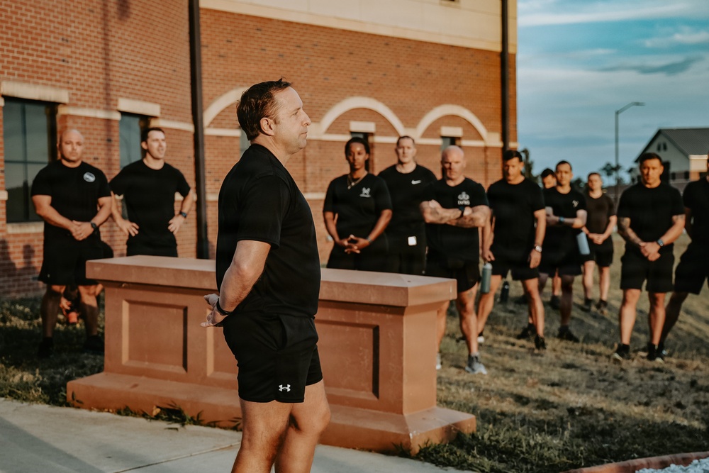 A group of approximately 15 individuals dressed in matching black athletic wear are assembled outdoors in front of a brick building, appearing to be in a formation or briefing. A man in the foreground wearing Under Armour black shorts and a black t-shirt appears to be addressing or leading the group. The setting and uniformity of attire suggest a military, law enforcement, or organized fitness training context.