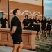 A group of approximately 15 individuals dressed in matching black athletic wear are assembled outdoors in front of a brick building, appearing to be in a formation or briefing. A man in the foreground wearing Under Armour black shorts and a black t-shirt appears to be addressing or leading the group. The setting and uniformity of attire suggest a military, law enforcement, or organized fitness training context.