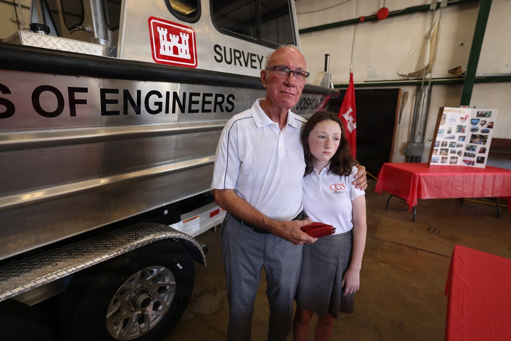 An older man and a young girl stand together in front of a U.S. Army Corps of Engineers survey vessel inside what appears to be a hangar or large indoor facility. The man has his arm around the girl in what appears to be a solemn or formal occasion. In the background, red-draped tables and a photo display board are visible, suggesting a commemorative or ceremonial event.