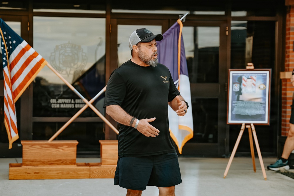 A middle-aged bearded man wearing a black t-shirt, black shorts, and a gray trucker cap appears to be speaking or presenting outdoors in front of a building entrance. Behind him are crossed American and purple/gold state flags mounted on wooden bases, along with a framed commemorative display on an easel. The setting appears to be a military or government facility based on visible signage referencing military ranks.