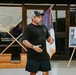 A middle-aged bearded man wearing a black t-shirt, black shorts, and a gray trucker cap appears to be speaking or presenting outdoors in front of a building entrance. Behind him are crossed American and purple/gold state flags mounted on wooden bases, along with a framed commemorative display on an easel. The setting appears to be a military or government facility based on visible signage referencing military ranks.