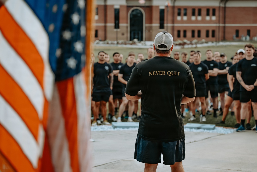 A leader or instructor wearing a black 'NEVER QUIT' t-shirt and white baseball cap stands with his back to the camera, addressing a large group of individuals dressed in matching black athletic wear. An American flag is prominently visible in the foreground left. The setting appears to be an outdoor formation or assembly in front of a brick institutional building.