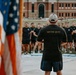 A leader or instructor wearing a black 'NEVER QUIT' t-shirt and white baseball cap stands with his back to the camera, addressing a large group of individuals dressed in matching black athletic wear. An American flag is prominently visible in the foreground left. The setting appears to be an outdoor formation or assembly in front of a brick institutional building.