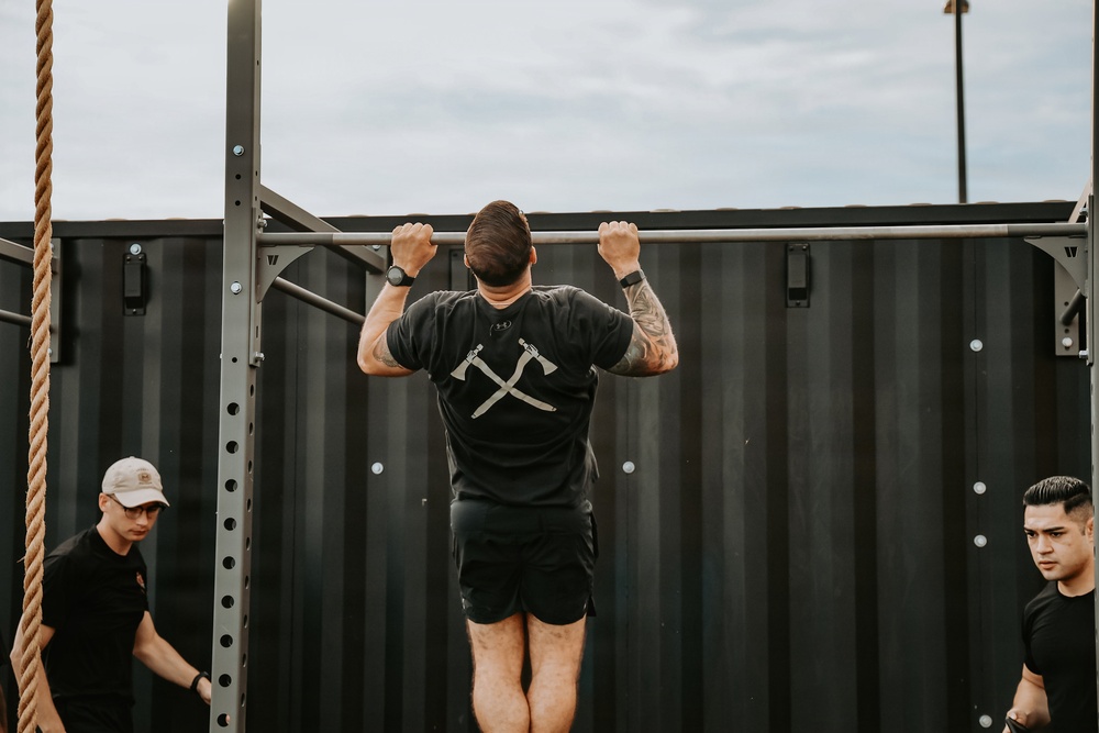 A man is performing pull-ups on an outdoor metal pull-up bar rig, with his back facing the camera. He is wearing a black Under Armour t-shirt featuring a crossed axe and sword graphic, black shorts, and has visible tattoos on both arms. Two other individuals dressed in black are present on either side of him, appearing to observe or wait their turn.