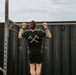 A man is performing pull-ups on an outdoor metal pull-up bar rig, with his back facing the camera. He is wearing a black Under Armour t-shirt featuring a crossed axe and sword graphic, black shorts, and has visible tattoos on both arms. Two other individuals dressed in black are present on either side of him, appearing to observe or wait their turn.