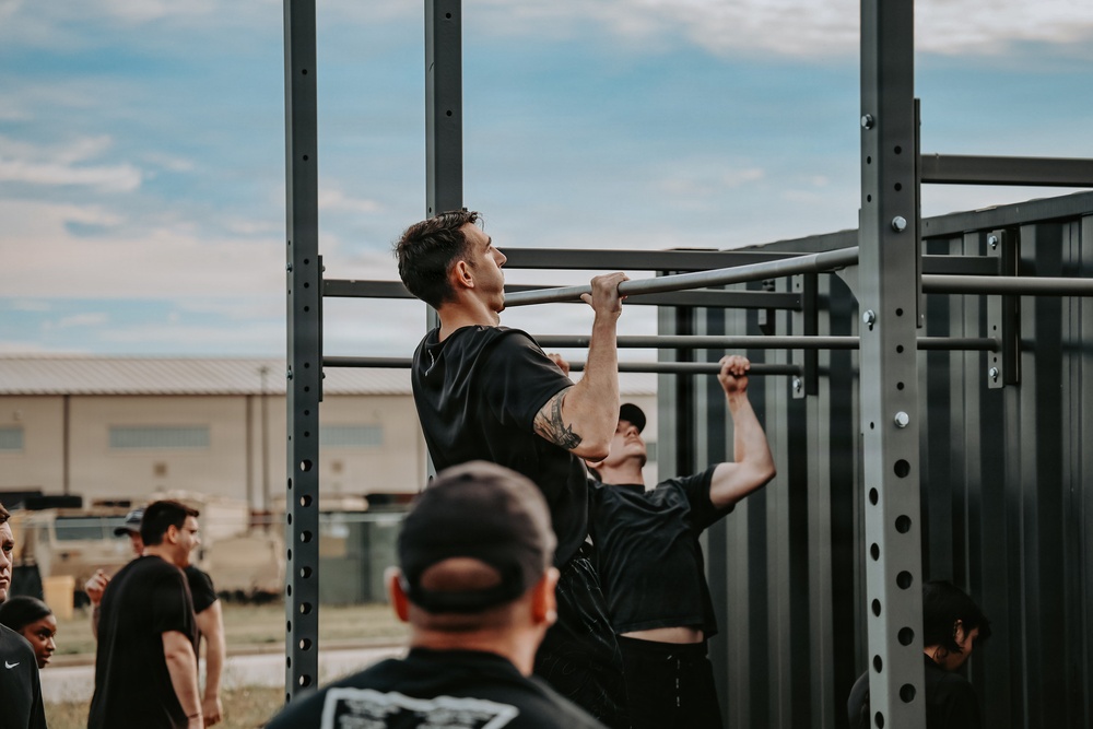 An outdoor fitness event or training session where multiple individuals are performing pull-ups on a large metal pull-up rig structure. A tattooed man in a black t-shirt is prominently featured at the top of a pull-up, with at least one other person visible doing the same exercise beside him. A group of observers dressed in black clothing watch from the surrounding area.