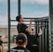 An outdoor fitness event or training session where multiple individuals are performing pull-ups on a large metal pull-up rig structure. A tattooed man in a black t-shirt is prominently featured at the top of a pull-up, with at least one other person visible doing the same exercise beside him. A group of observers dressed in black clothing watch from the surrounding area.