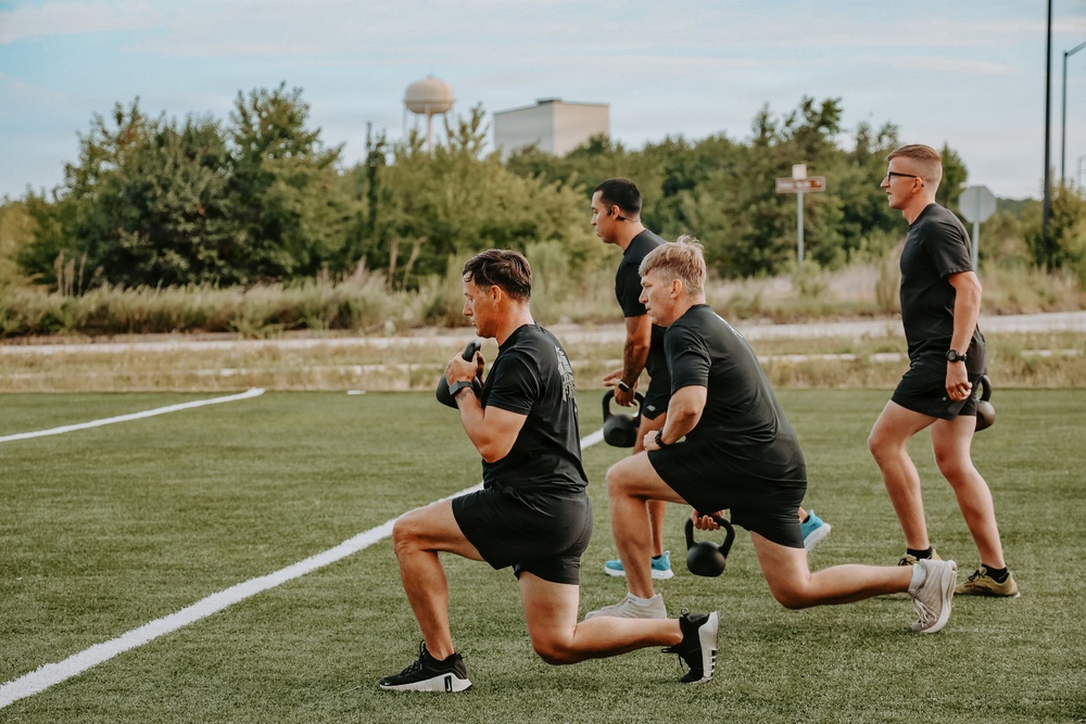 Four men dressed in matching black t-shirts and shorts are performing kettlebell lunges on an outdoor artificial turf field. They appear to be participating in a group fitness training session, all executing the same exercise in synchronized form. The setting appears to be a sports field with trees, a water tower, and a building visible in the background.