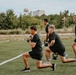 Four men dressed in matching black t-shirts and shorts are performing kettlebell lunges on an outdoor artificial turf field. They appear to be participating in a group fitness training session, all executing the same exercise in synchronized form. The setting appears to be a sports field with trees, a water tower, and a building visible in the background.