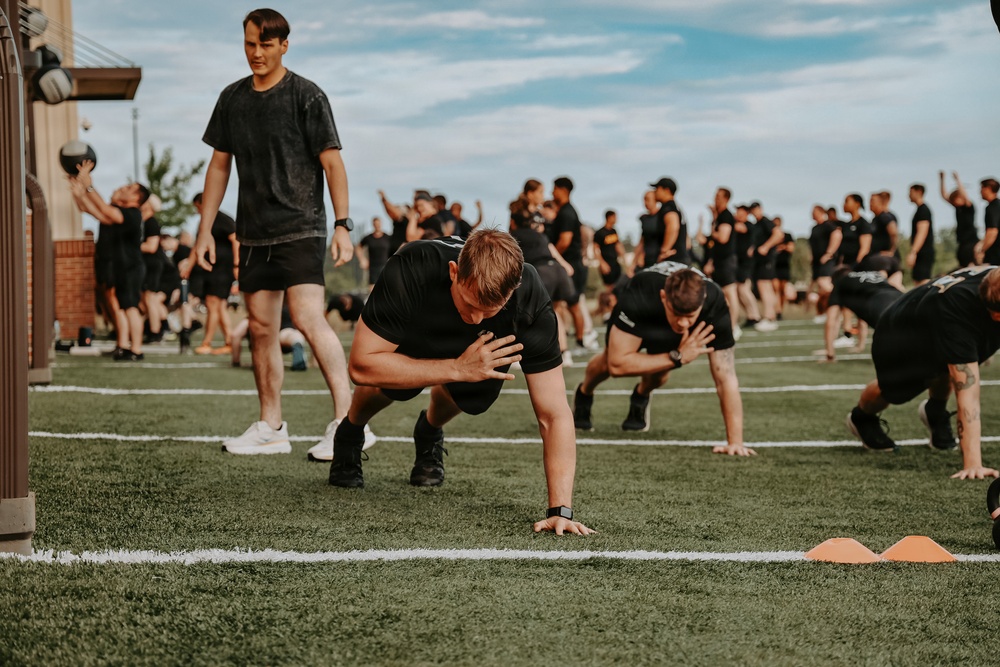 A large group of individuals dressed in matching black t-shirts and shorts are participating in an organized physical training session on an artificial turf field. Several participants in the foreground are performing push-ups or a push-up variation, while one standing individual appears to be an instructor or supervisor holding a medicine ball. The background shows dozens more participants engaged in various exercises, suggesting a structured group fitness or military-style PT event.